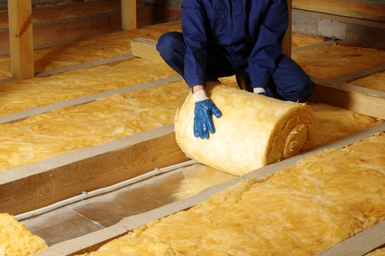 Ouvrier en combinaison bleue installant isolation thermique dans combles, manipulant rouleau de laine min&eacute;rale jaune entre solives bois