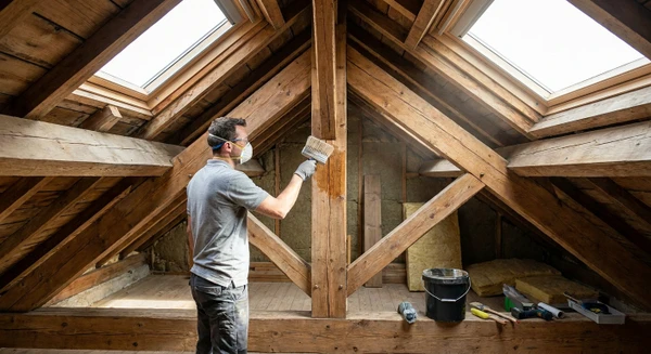 Photographie professionnelle montrant un artisan qualifi&eacute; dans les combles ou sous la toiture d'une maison, traitant les &eacute;l&eacute;ments structurels de la charpente (poutres, solives) avec un produit de protection/traitement anti-xylophages. L'artisan porte des &eacute;quipements de protection appropri&eacute;s (masque, gants), utilise un &eacute;quipement de traitement professionnel (pulv&eacute;risateur ou pinceau de traitement sp&eacute;cialis&eacute;), et travaille sur les bois de structure visibles. La lumi&egrave;re naturelle provenant des ouvertures de toiture &eacute;claire la sc&egrave;ne et met en &eacute;vidence le travail de traitement en cours.