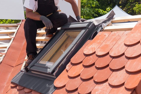 Ouvrier en salopette noire installant une fen&ecirc;tre de toit sur une toiture en tuiles terre cuite rouge