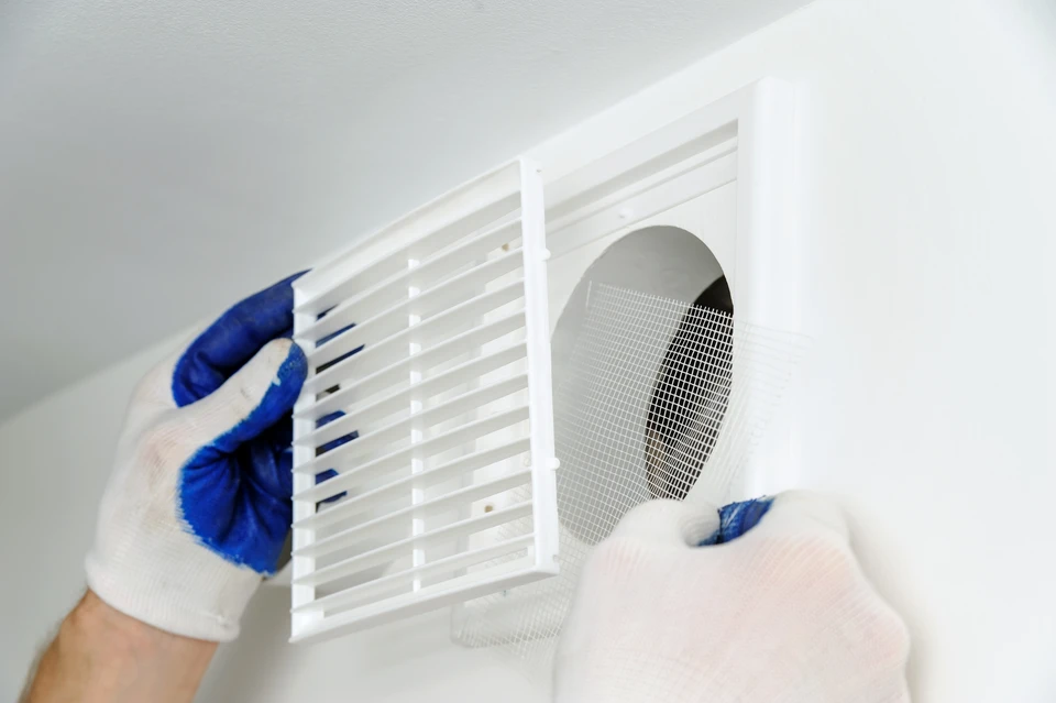 Intérieur résidentiel, technicien en gants de protection bleus installe une grille d'aération blanche sur conduit de ventilation mural